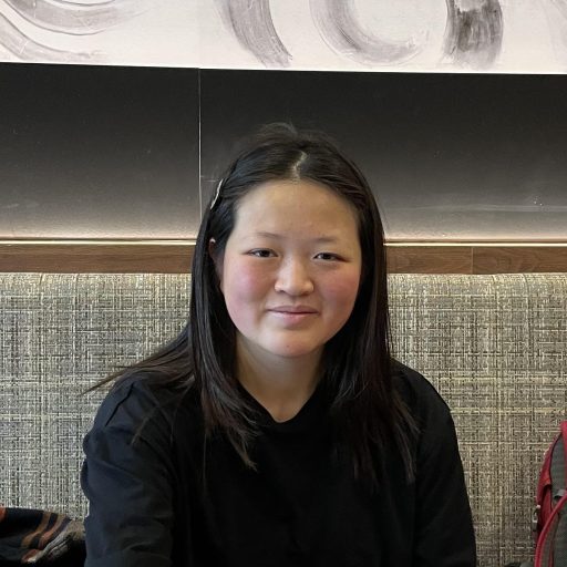 Person seated indoors, facing the camera with a calm smile, wearing a black top, with shoulder-length dark hair, against a neutral wall and upholstered bench background.